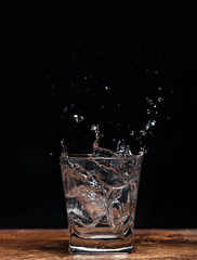 A glass tumbler with splashes on a dark background and a wooden table into which ice cubes had fallen