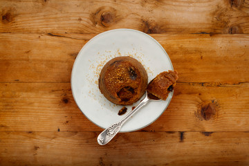 An individual spotted dick sponge cake, on a white plate with a spoon and sprinckled brown sugar,...