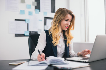 young blond beautiful woman manager works in her laptop in modern office, taking notes to her business planner,looks confident and busy, multitasking, work concept