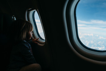 Caucasian little girl traveling by an airplane. Child sitting by aircraft window and looking to the sky anf plane wing. Concept of traveling with kids