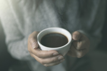 Closeup of female hands with a cup of hot coffee. in the morning soft sunlight. Beautiful girl in grey sweater.