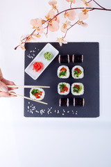 Sushi set with ginger and wassabi on a black square plate of stone on a light background. Near a sprig of sakura and female hand holds sushi with chopsticks.. Top view