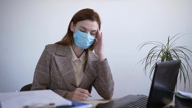 feeling unwell, a young female bises employee works in a medical protective mask against infection and coronavirus works at a laptop computer and suffers from severe headache, holds whiskey with her