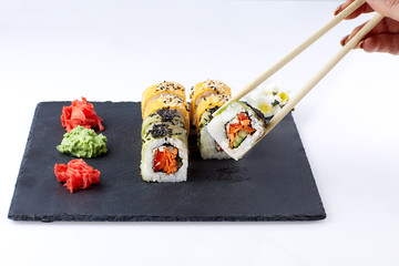 Sushi set with ginger and wassabi on a black square plate of stone on a light background. Female hand holds sushi with chopsticks.