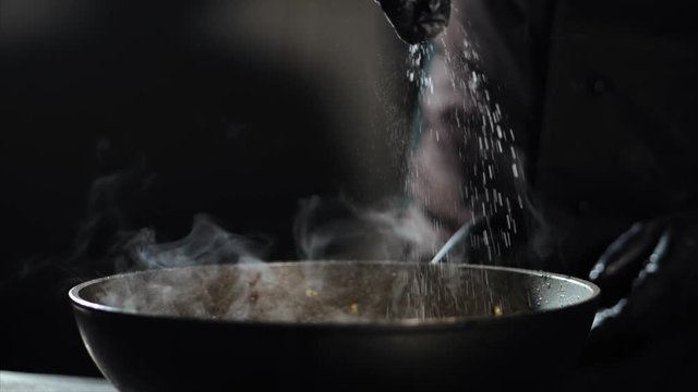 Chef is cooking in wok pan at commercial kitchen. Cook is salting food while preparing it in wok pan in slow motion.