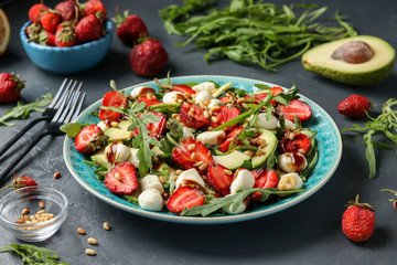 Healthy salad with strawberries, avocado, arugula and mozzarella, dressed with olive oil and balsamic dressing in a blue plate on a dark background, Closeup