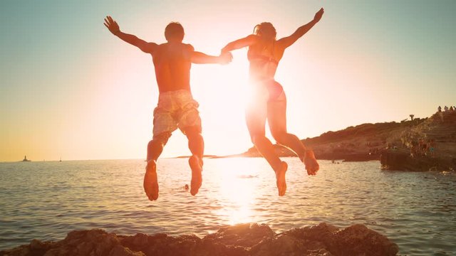 SLOW MOTION, LOW ANGLE, LENS FLARE: Cheerful couple on summer vacation jumps into the ocean at sunrise. Golden evening sun rays shine on young man holding girlfriend's hand as they jump in the water.