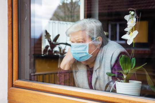 Senior woman with surgical mask sitting on a window at home, coronavirus and covid-19 provisions