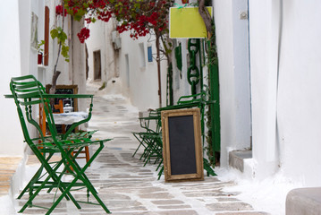 Fototapeta na wymiar A narrow alleyway in the old town, at the beautiful Greek island of Ios. Picture of a charming small cafe with traditional chairs and tables in the shade of bougainvillea.