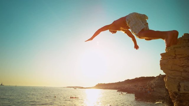 SLOW MOTION, LENS FLARE, CLOSE UP: Athletic man dives off a cliff and head-first into the ocean at sunset. Fit male tourist jumps of a rocky ledge and dives into the refreshing sea water in Croatia.