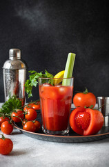 Classic alcoholic cocktail Bloody Mary with ice, lemon and celery. Cocktail ingredients and bar tools on a gray table and black background. Vertical 