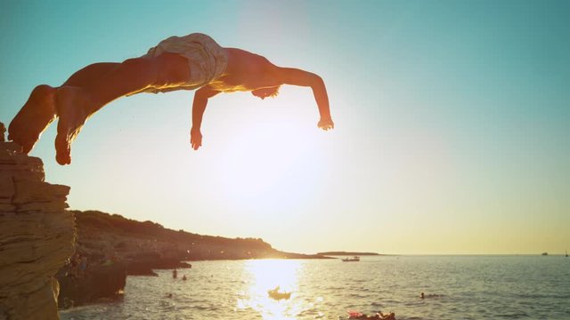 SLOW MOTION, CLOSE UP, LENS FLARE: Fit male tourist dives into the refreshing ocean water at sunset. Athletic man on summer vacation in Croatia dives off a rocky cliff on a perfect summer evening.