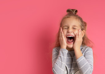 Little blonde female with bun hairstyle, dressed in gray striped blouse. She laughing out loudly, touching cheeks, posing against pink background. Childhood, fashion, advertising. Close up, copy space