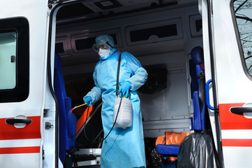 Paramedic in protective mask and costume disinfecting the ambulance car with sprayer