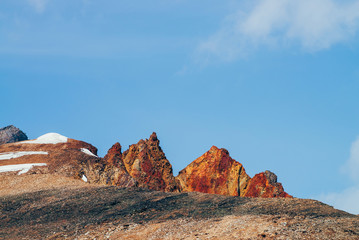Fototapeta premium Awesome sunny mountain with red orange brown crags in top under blue sky. Colorful stony hill with snow in sunshine. Wonderful alpine scenery with rock in red color. Beautiful vivid highland landscape