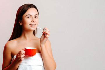 Young woman after shower enjoying coffee and making makeup in white towel and bare shoulders. Concept of multitasking. Girl is drinking coffe and putting on lipstick.