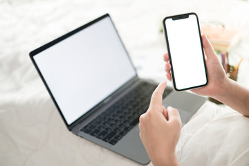 Young woman hand holding blank screen phone and blank screen phone and laptop computer in the bed room, Work form home and holiday concept 
