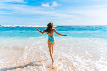 A girl runs along the beach on the water in the blue waves of the ocean. Carefree holidays on the Sunny beaches of Bali. Melasti beach in Bali.
