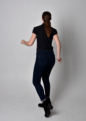  Portrait of a pretty brunette girl with ponytail, wearing jeans boots and a black blouse.  full length standing pose, with back to the camera on a studio background.