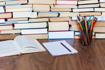 Open textbook, notebook, glasses, pencils in holder, stack of old book on wooden table, education concept background, many books piles with copy space for text.