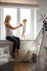 Portrait of a smiling young cute mother and her son playing near the window in a bright white room. Sunny day outside. True mother love