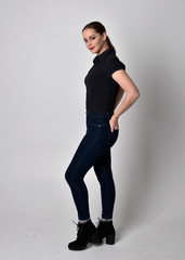 Portrait of a pretty brunette girl with ponytail, wearing jeans boots and a black blouse.  full length standing pose  in side profile on a studio background.