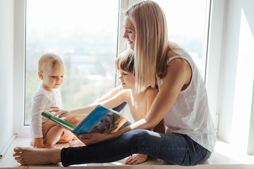Portrait of a smiling young cute mother and her sons reading a book near the window in a bright white room. Sunny day outside. 