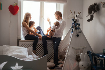 Happy smiling modern caucasian family playing and spending time in a bright white room near the window. Sunny day. Perfect parenting relationship. Parents of two cute boys.