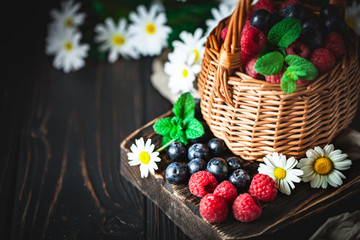 Raspberries and blueberries in a basket with chamomile and leaves on a dark background. Summer and healthy food concept. Background with copy space. Selective focus.
