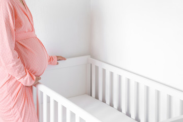 One young pregnant woman in pink robe standing beside white crib for future baby. Waiting concept....