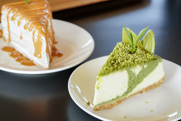 Sweet or dessert, a matcha green tea cheesecake topping with crush chocolate and decor with tea leaf on white plate on black table with a cafe environment and Thai milk tea Crepe Cake beside it