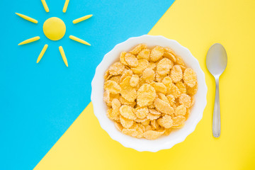 White bowl of corn flakes with spoon. Yellow sun shape on bright blue table. Simple daily healthy...