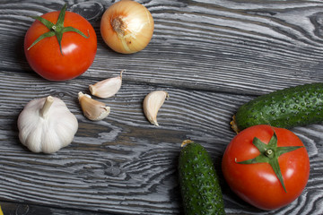 Tomatoes with green ponytails, onion and garlic head. Two green prickly cucumbers with yellow flowers. They lie on the surface of brushed pine boards.