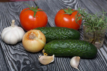 Tomatoes with green ponytails, onion, lemon and garlic head. Two green prickly cucumbers with yellow flowers. A glass with rosemary branches. They lie on the surface of brushed pine boards.