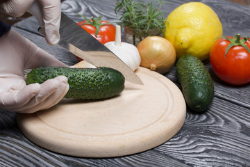A man in rubber gloves cuts a cucumber with a knife on a cutting board. Nearby are red tomatoes with green ponytails, rosemary and garlic. On the surface of brushed pine boards.