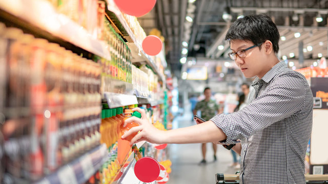 Asian Man Choosing Orange Juice In Supermarket Using Smartphone To Check Shopping List. Male Shopper With Shopping Cart Selecting Beverage Bottle Product In Grocery Store.