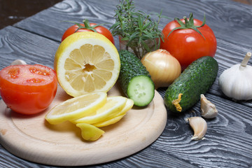 On a cutting board, sliced lemon, tomato and cucumber. Nearby are cucumber, rosemary, onions and garlic. On the surface of brushed pine boards.
