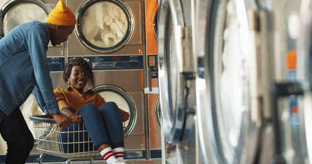 Obraz na płótnie Canvas Young African American cheerful couple having fun in laundry service room. Pretty happy girl sitting in carriage and guy riding her at working washing machines while cleaning clothes.