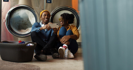 Fototapeta na wymiar African American young couple sitting on floor with basket of dirty clothes and watching video on smartphone while washing machines working Man and woman using phone and resting in public laundromat