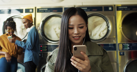 Asian pretty cheerful woman in headphones watching video on smartphone in laundry service room. Beautiful happy girl listening to music on phone and waiting for clothes to get clean in washhouse.