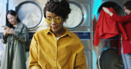 African American young pretty and stylish girl in yellow glasses standing in laundry service room and flipping pages of fashion journal. Woman reading magazine while waiting for clothes to be washed.