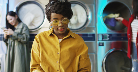 Fototapeta na wymiar African American young pretty and stylish girl in yellow glasses standing in laundry service room and flipping pages of fashion journal. Woman reading magazine while waiting for clothes to be washed.
