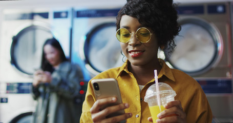 African American girl drinking orange juice with straw, texting message on phone and waiting for clothes to get clean Woman sipping drink in laundry service room and tapping or scrolling on smartphone