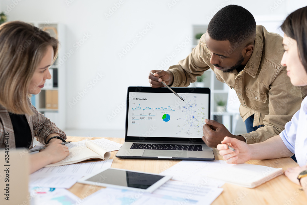 Poster afro-american marketer pointing at laptop screen while reporting marketing statistics at meeting
