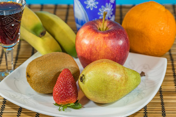 Fresh tropical and subtropical fruits in a large white dish. Cristal glass of red wine and blue and white carved crystal goblet on wicker bamboo stand on the dining table. Close up, Selective focus