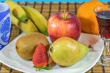 Fresh tropical and subtropical fruits in a large white dish. Cristal glass of red wine and blue and white carved crystal goblet on wicker bamboo stand on the dining table. Close up, Selective focus