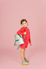 Cute little blonde classmate in a pink suit in the studio with a holographic backpack. Funny first grader on a pink pastel background with copyspace