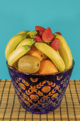Fresh tropical and subtropical fruits in a large blue crystal vase. Wicker bamboo stand on the dining table, blue background