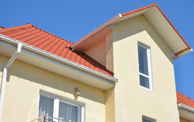 The painted facade of a two storey house with a gable roof covered with red metal roof tiles and a white roof gutter.
