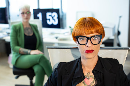 manager tatuata con i capelli rossi e gli occhi chiari egli occhiali ,indossa un completo nero nel suo ufficio con la colega sullo sfondo